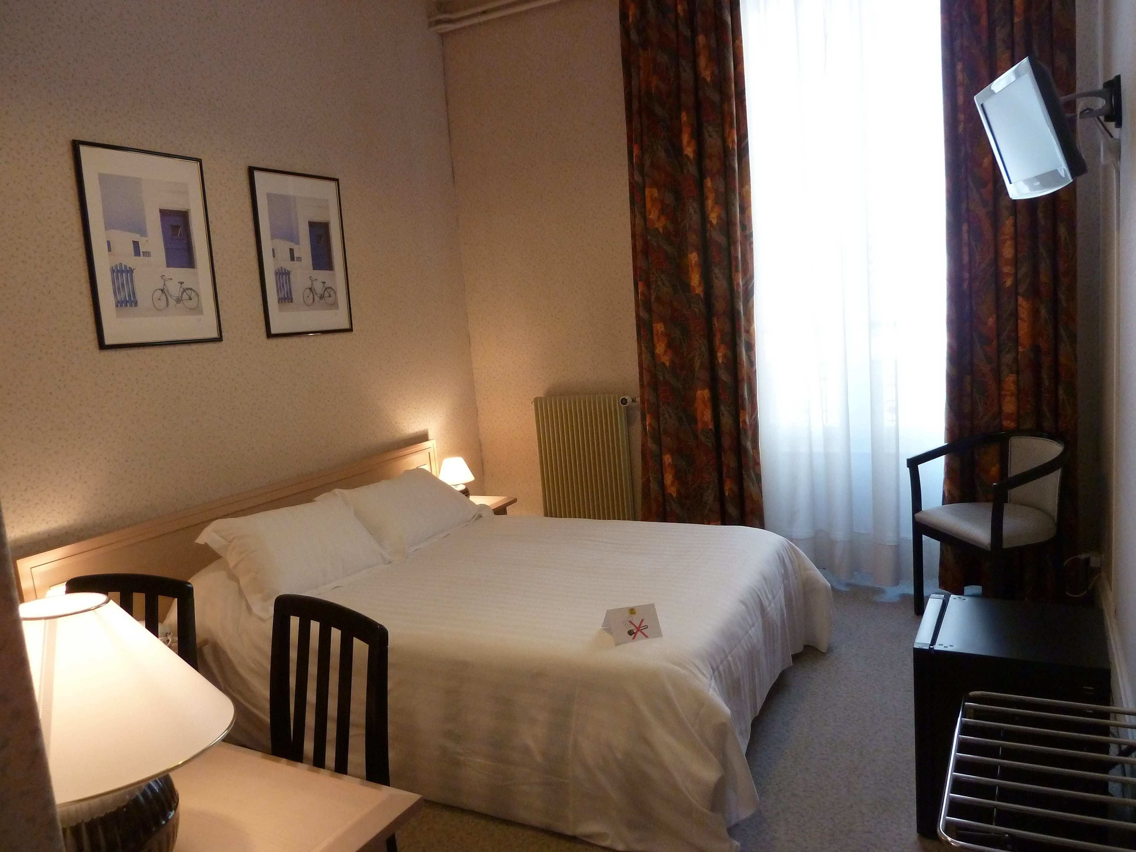A hotel room with a bed, a desk with a lamp, a chair, a TV mounted on the wall, and two framed pictures on the wall.