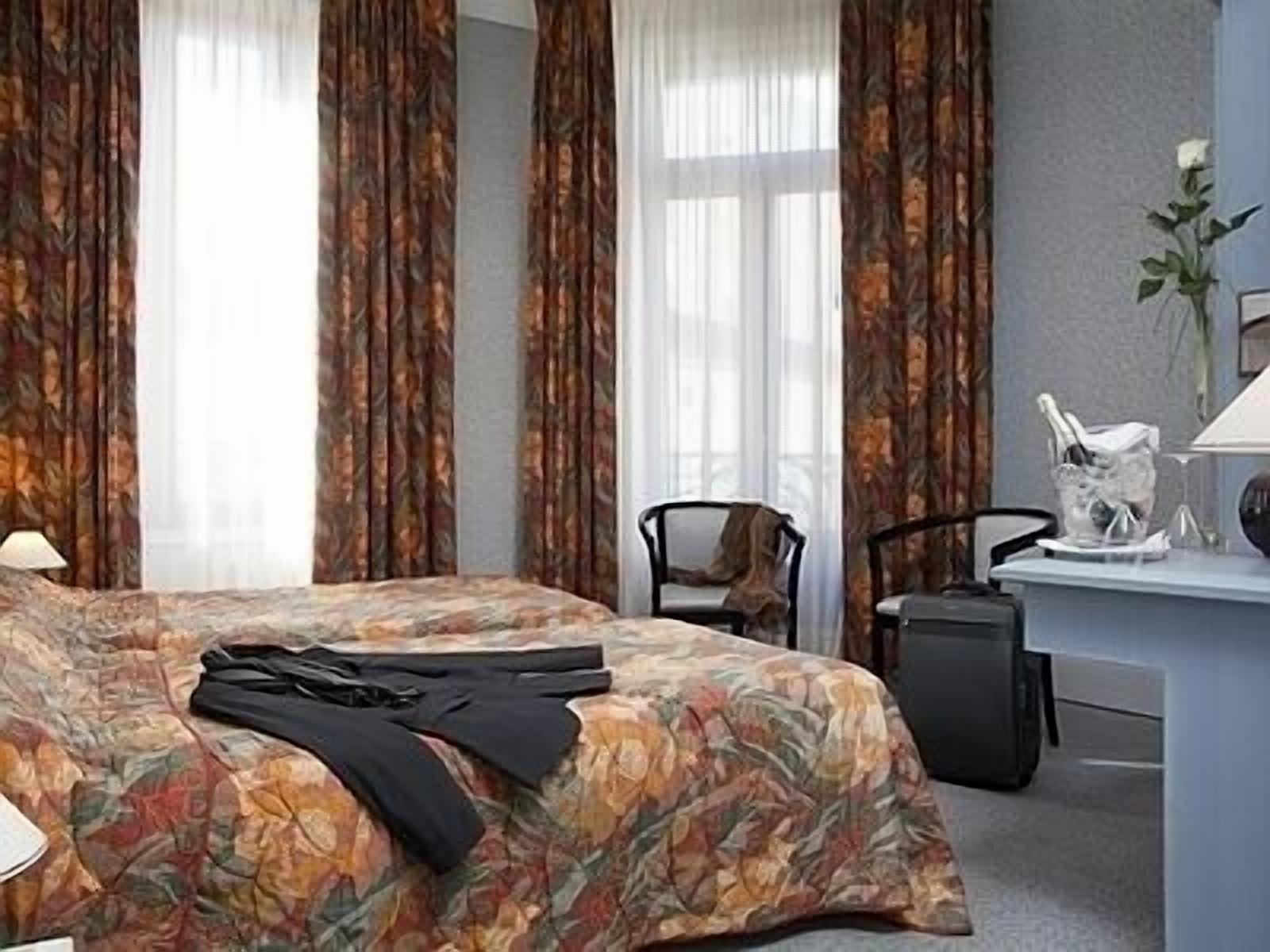 Habitación de hotel con cubrecama floral, un escritorio con lámpara, una silla y una pequeña planta.