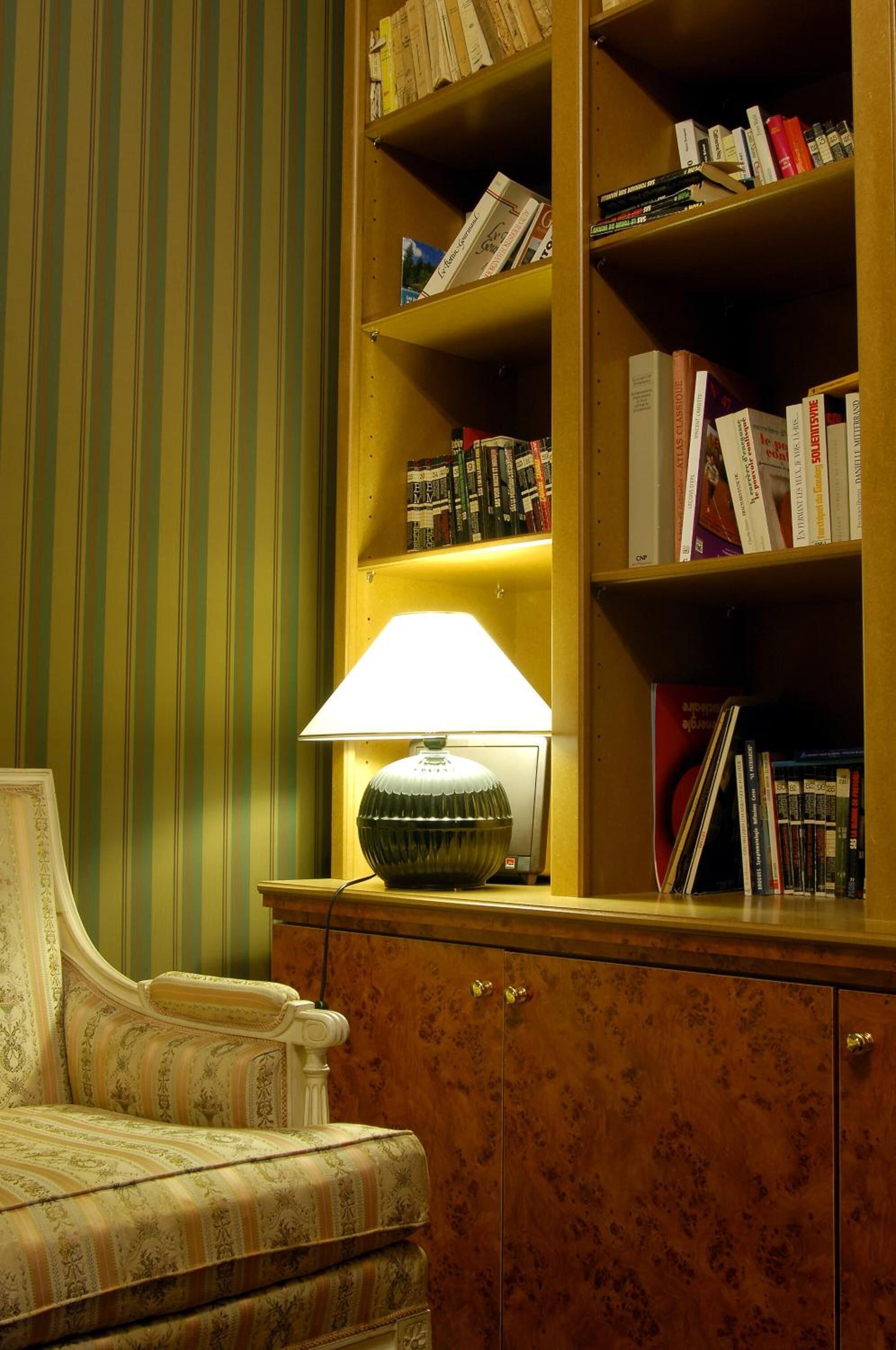 Een kamer met gestreepte behang, een houten boekenkast, een lamp en een fauteuil met een patroon.