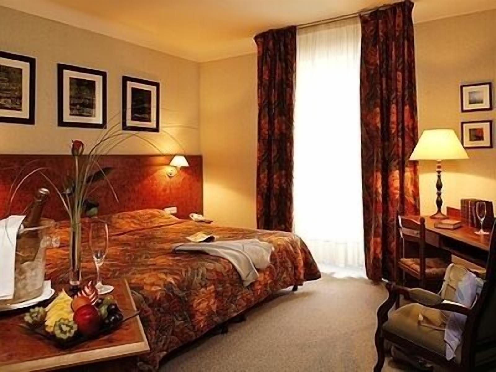 Une chambre d’hôtel avec un lit, un bureau doté d’une lampe, une chaise et une table à manger agrémentée de fruits et d’un vase.