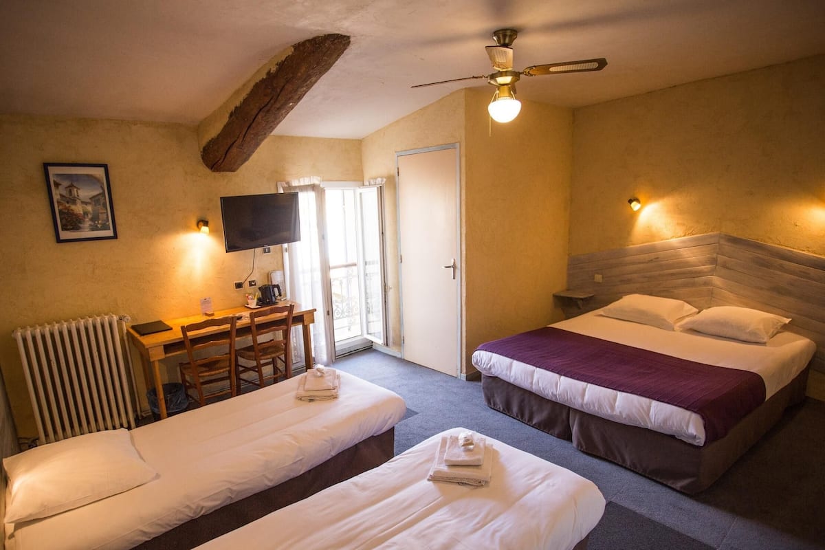 A hotel room with two beds, a desk, a chair, a television, and a ceiling fan.