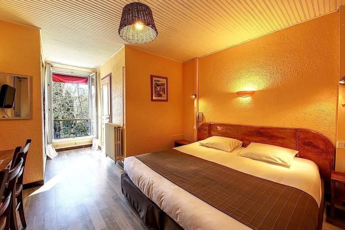 A bedroom with a large bed, a wooden headboard, a bedside table, a hanging light fixture, and a view of the outdoors through a door.