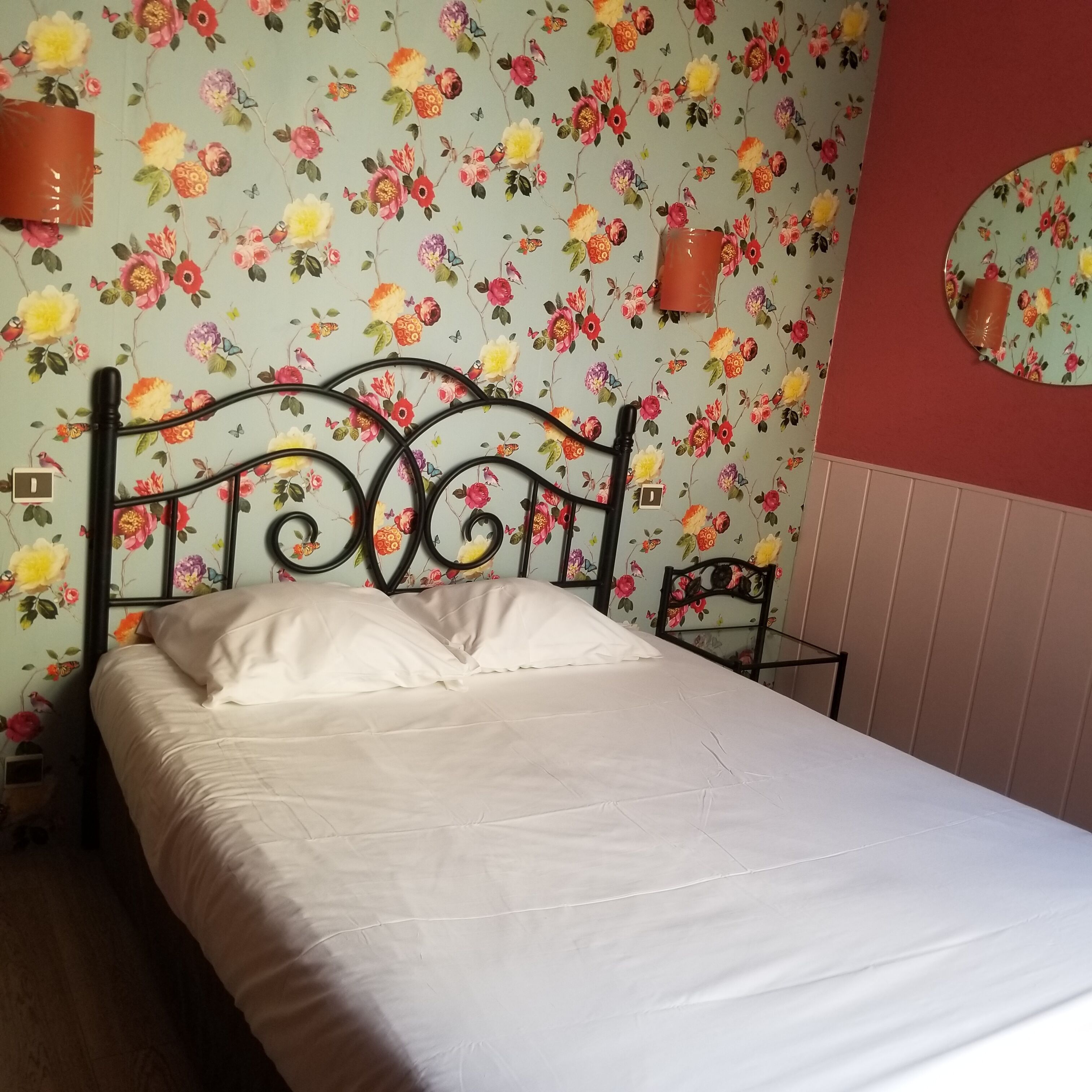 A bed with a floral wallpaper background, a round mirror on the wall, and a small table with a chair.