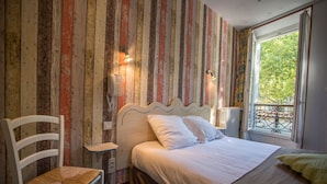 Un dormitorio con pared a rayas, una cama con ropa blanca, una silla de madera y una ventana con vistas a árboles.