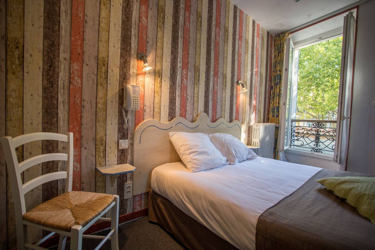 A bedroom with a striped wall, a bed with white bedding, a wooden chair, and a window with a view of trees.