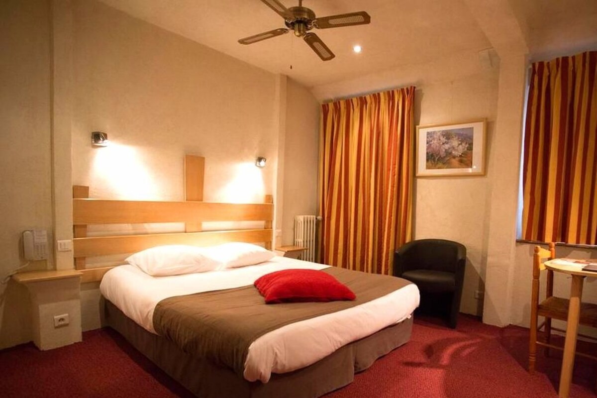 A hotel room with a bed, a desk, a chair, a ceiling fan, and curtains.