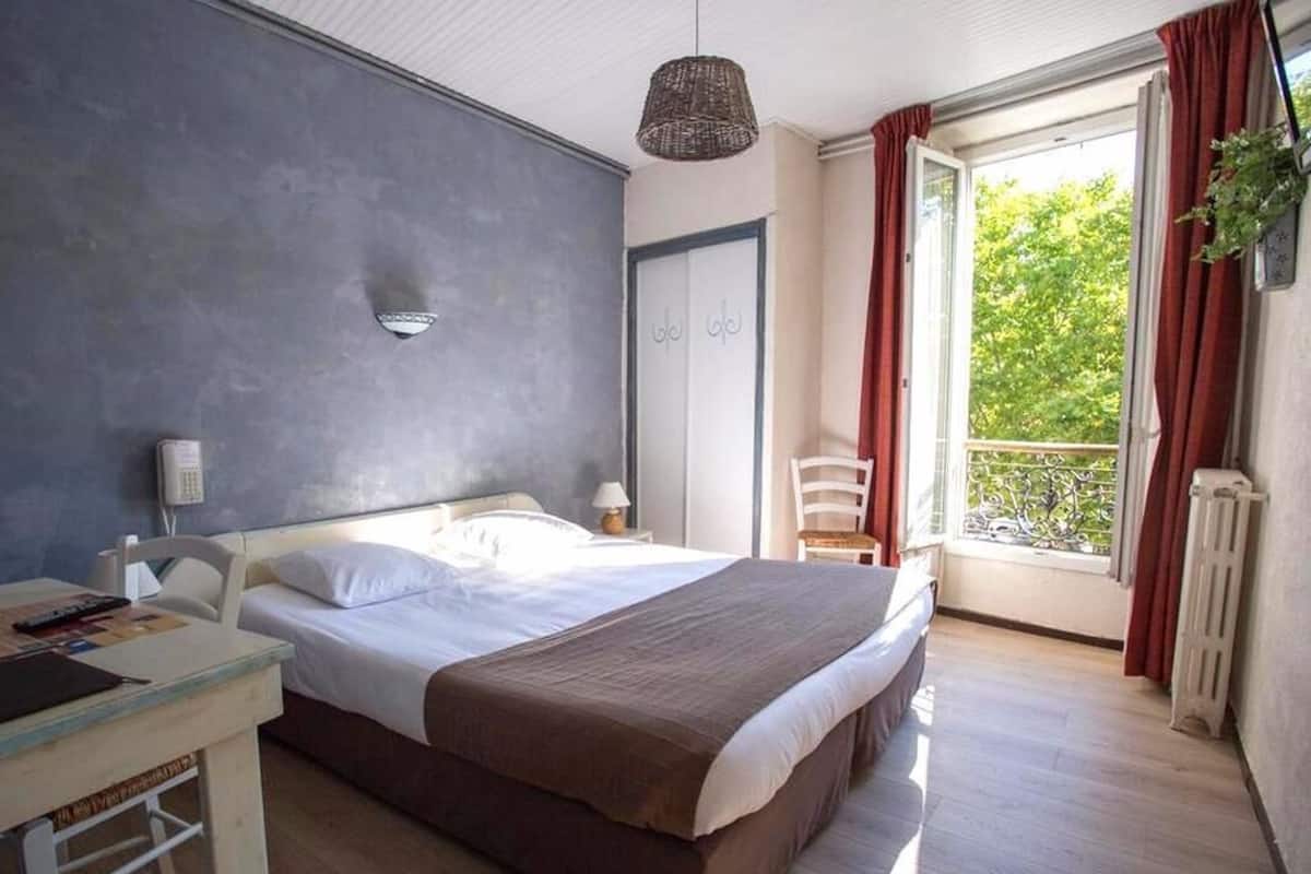 A bedroom with a large bed, a bedside table, a chair, a hanging plant, and a window with curtains.