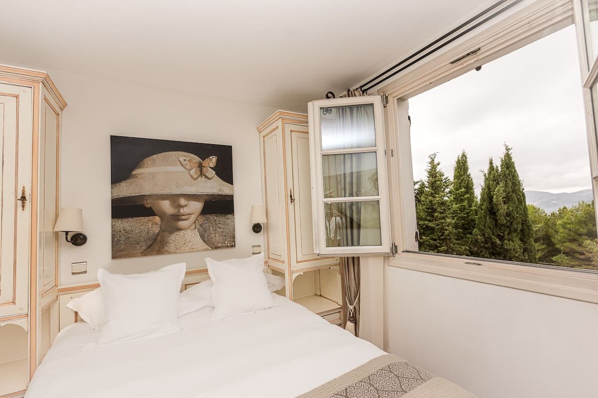 A bedroom with a bed, a painting, a window with curtains, and a view of trees.