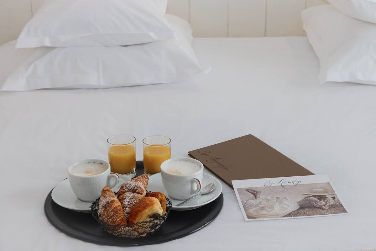 A neatly made bed with white linens, a breakfast tray with croissants, two cups of coffee, and two glasses of orange juice.