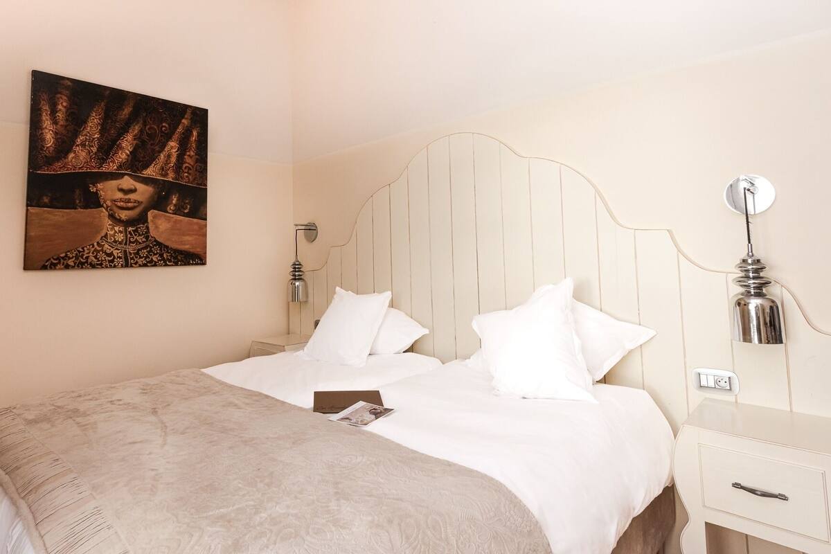 A neatly made bed with white linens and a beige blanket, a bedside table with a lamp, and a wall-mounted light fixture.
