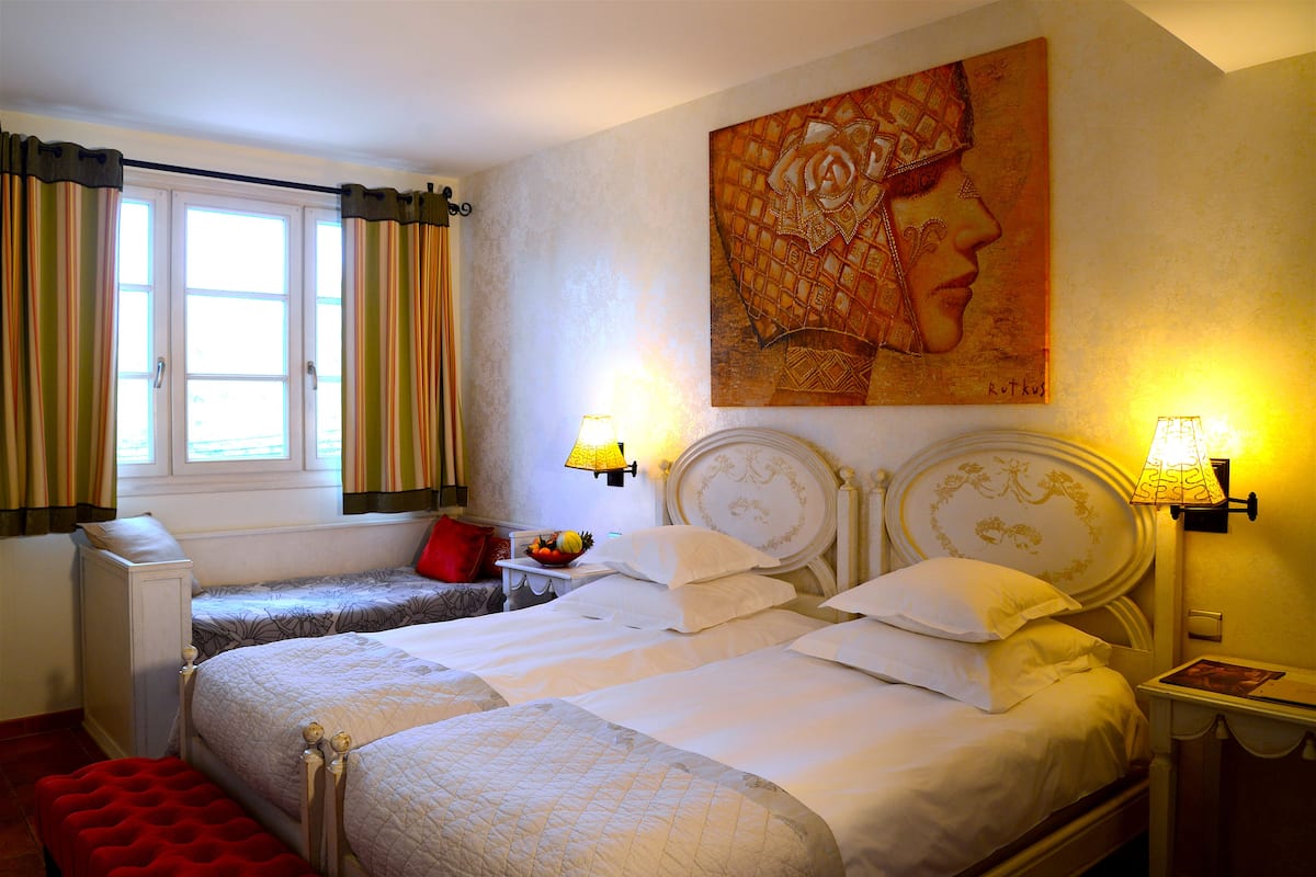 A double bed with a red bench, a bedside table with a fruit bowl, and a painting on the wall.