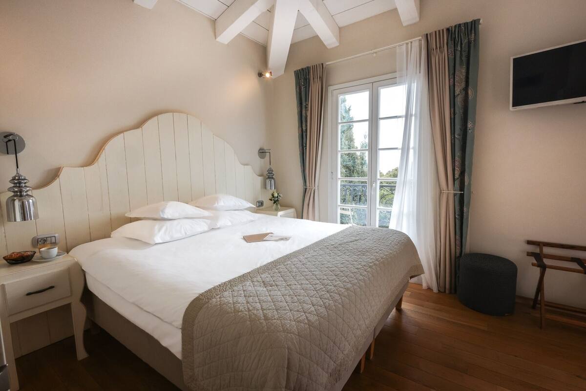 A bedroom with a large bed, a bedside table with a lamp, a small table with a cup and a pastry, and a TV on the wall.