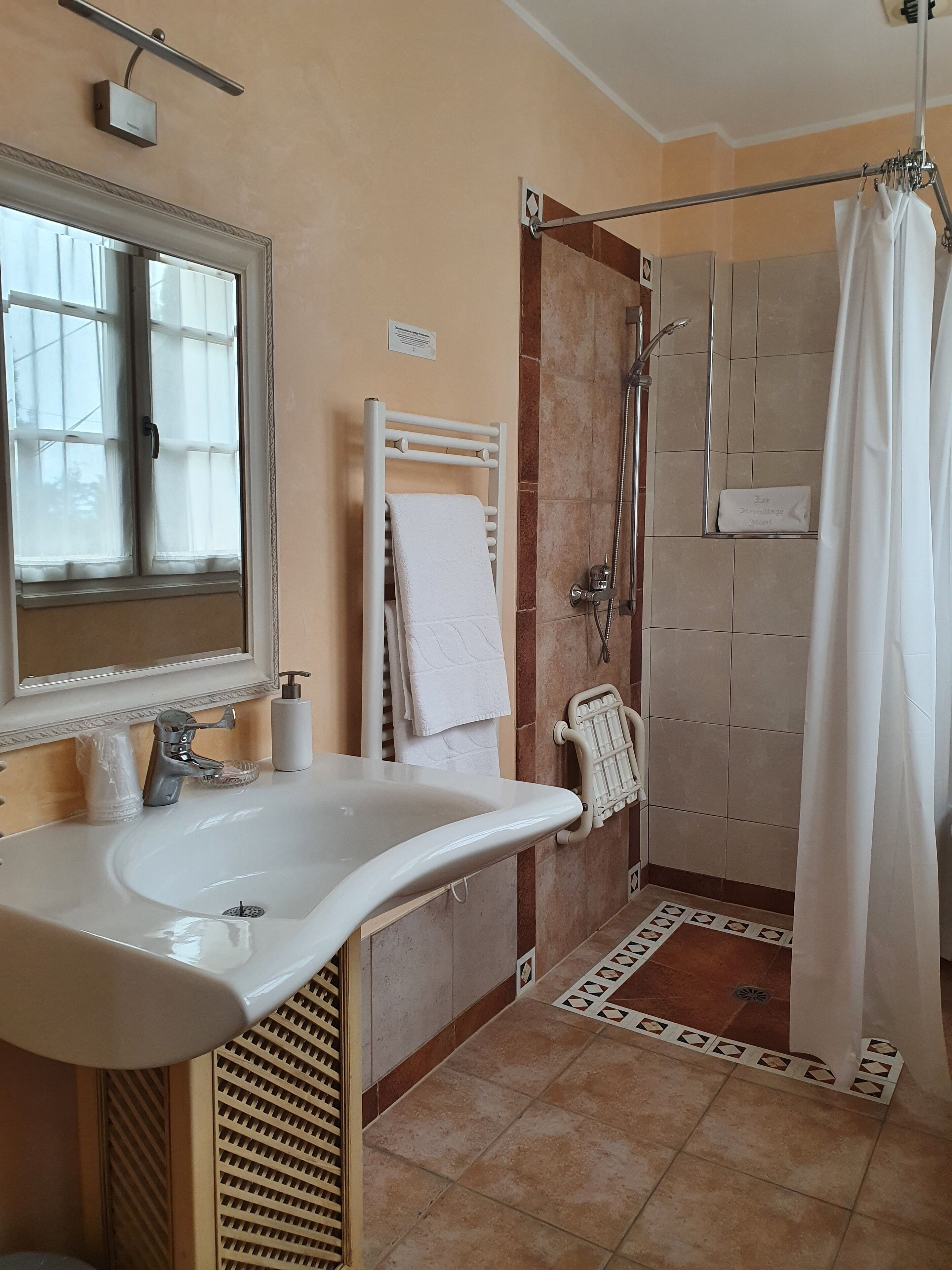 Ein Badezimmer mit einem weißen Waschbecken, einem Spiegel, einem Handtuchhalter, einer Dusche und einem gefliesten Boden.