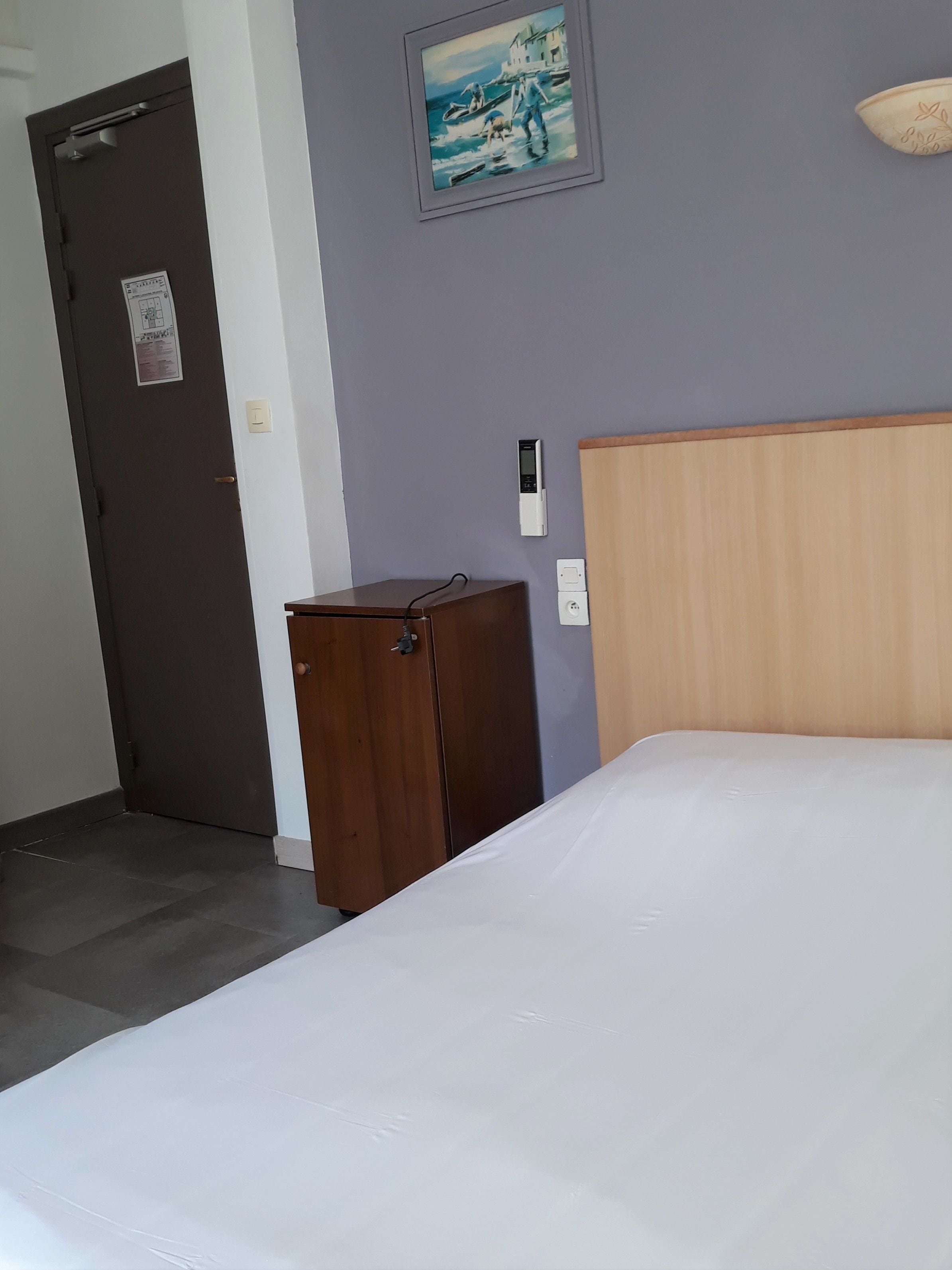 Une chambre d’hôtel avec un lit, une tête de lit en bois, une petite armoire en bois, un tableau accroché au mur et une porte avec une pancarte.