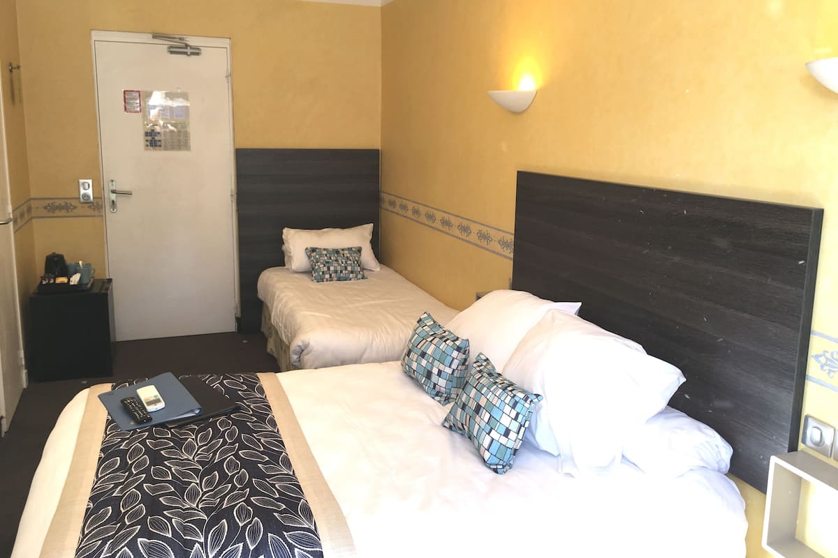 A hotel room with two beds, a bedside table, a nightstand, an air conditioning unit, and a wall-mounted light fixture.