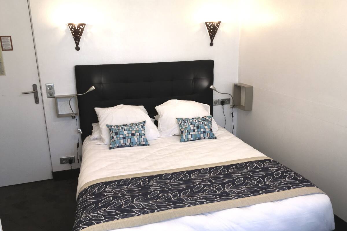 A neatly made bed with a black headboard, white pillows, and a patterned bedspread. There are two wall-mounted light fixtures on either side of the bed.
