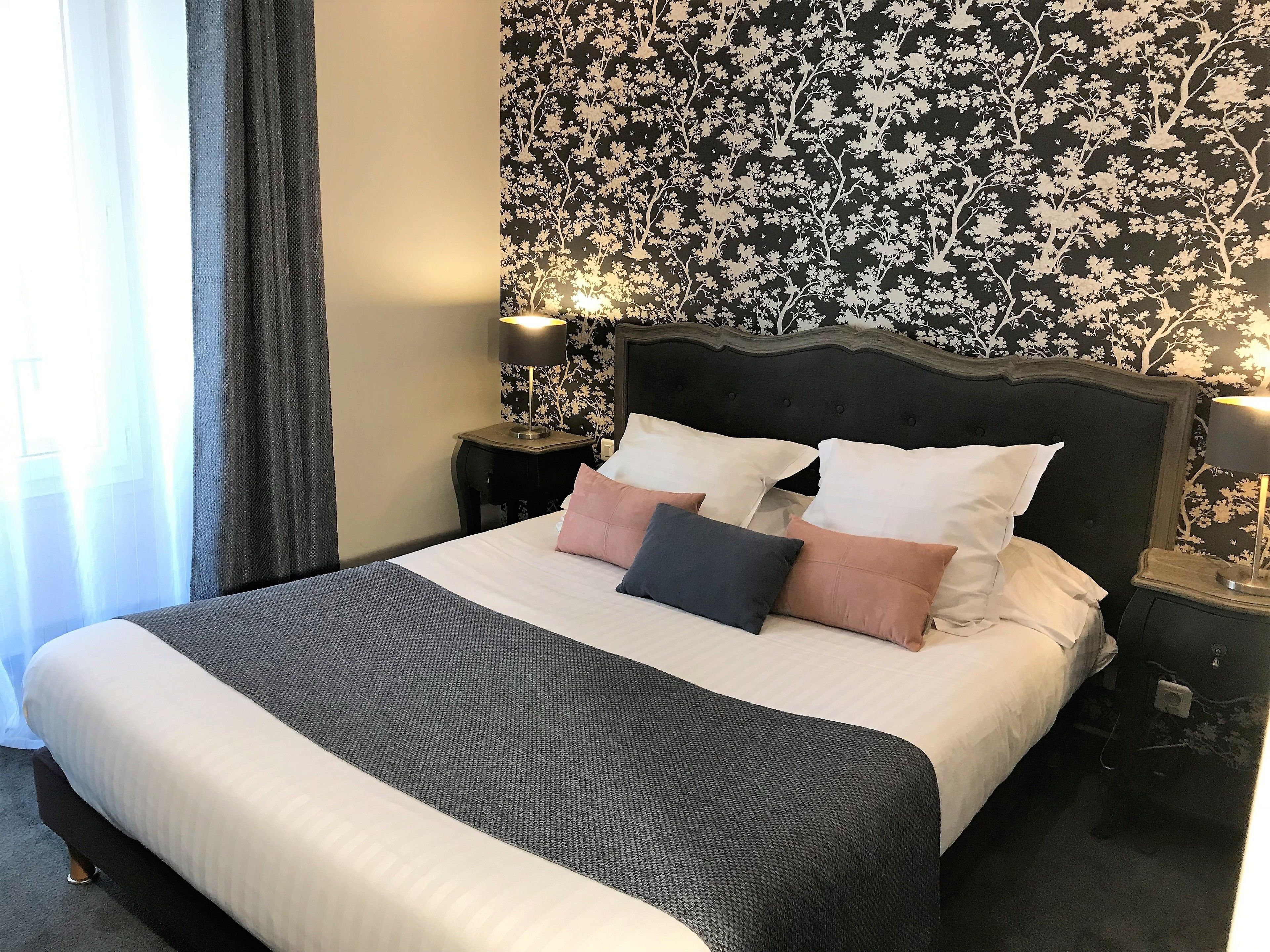 A neatly made bed with a dark headboard and multiple pillows, a small bedside table with a lamp, and a floral wallpaper pattern on the wall.