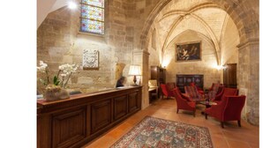 A room with a stone wall, a wooden reception desk, red upholstered chairs, and a patterned rug. - Hotel des Augustins (Aix-en-Provence)