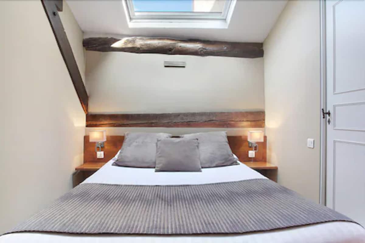 A bedroom with a sloped ceiling, exposed wooden beams, a bed with a grey blanket, two bedside lamps, and a skylight.