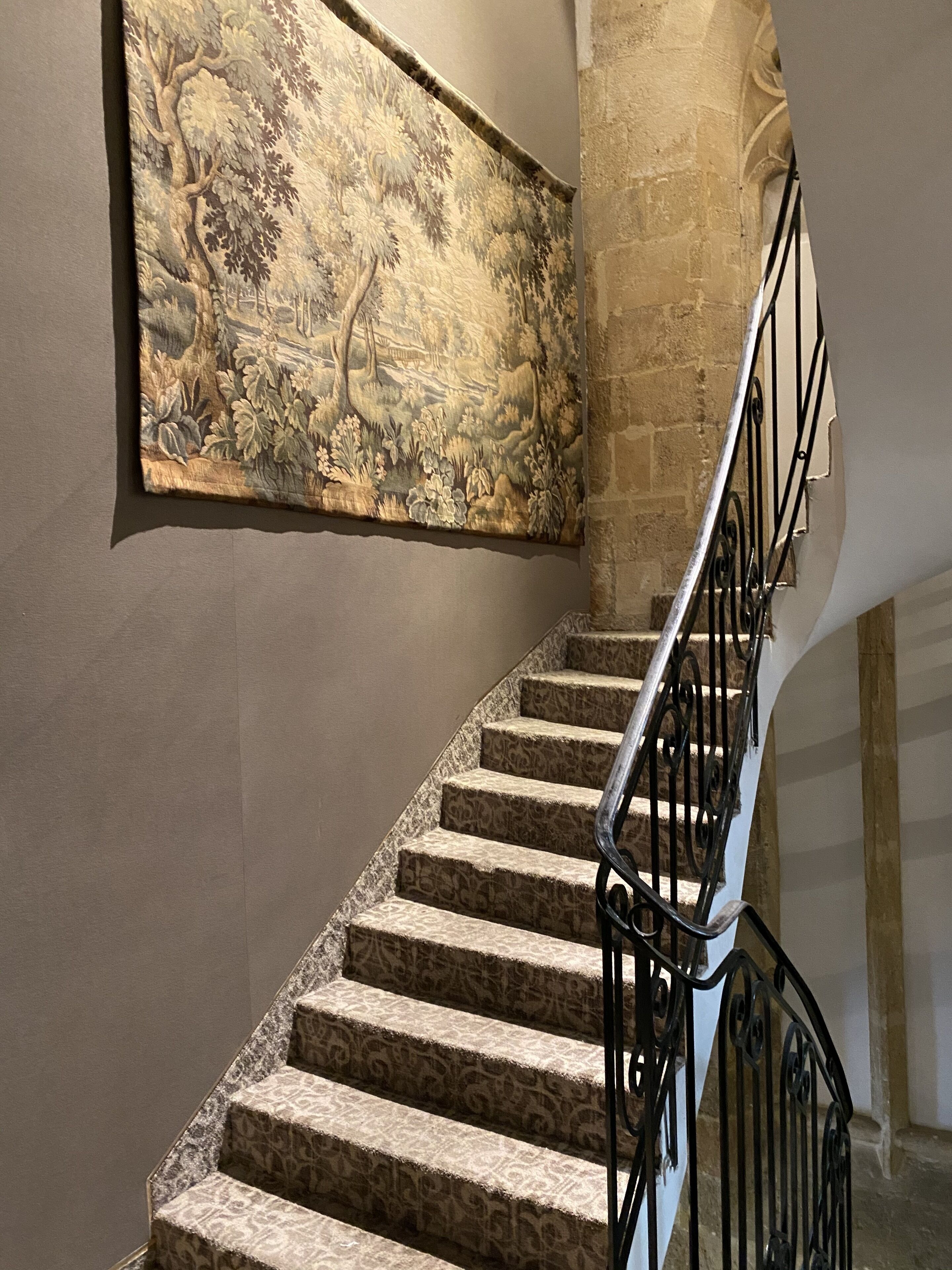 Un escalier aux marches décorées et une rampe en métal, menant à un mur de pierre orné d’une grande tapisserie florale.
