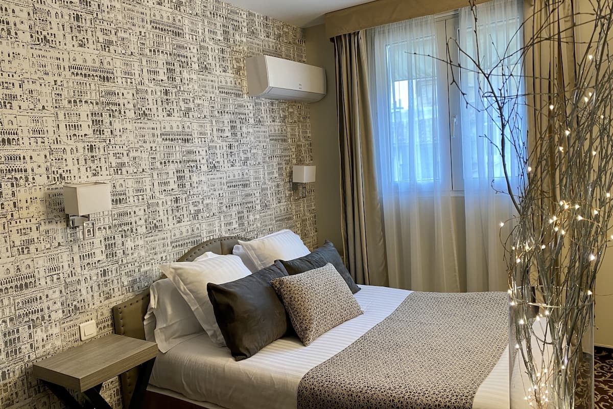 A hotel room with a patterned wallpaper, a bed with multiple pillows, a bedside table, an air conditioning unit, a window with curtains, and a decorative branch with lights.