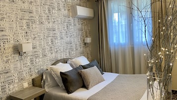 A hotel room with a patterned wallpaper, a bed with multiple pillows, a bedside table, an air conditioning unit, a window with curtains, and a decorative branch with lights.