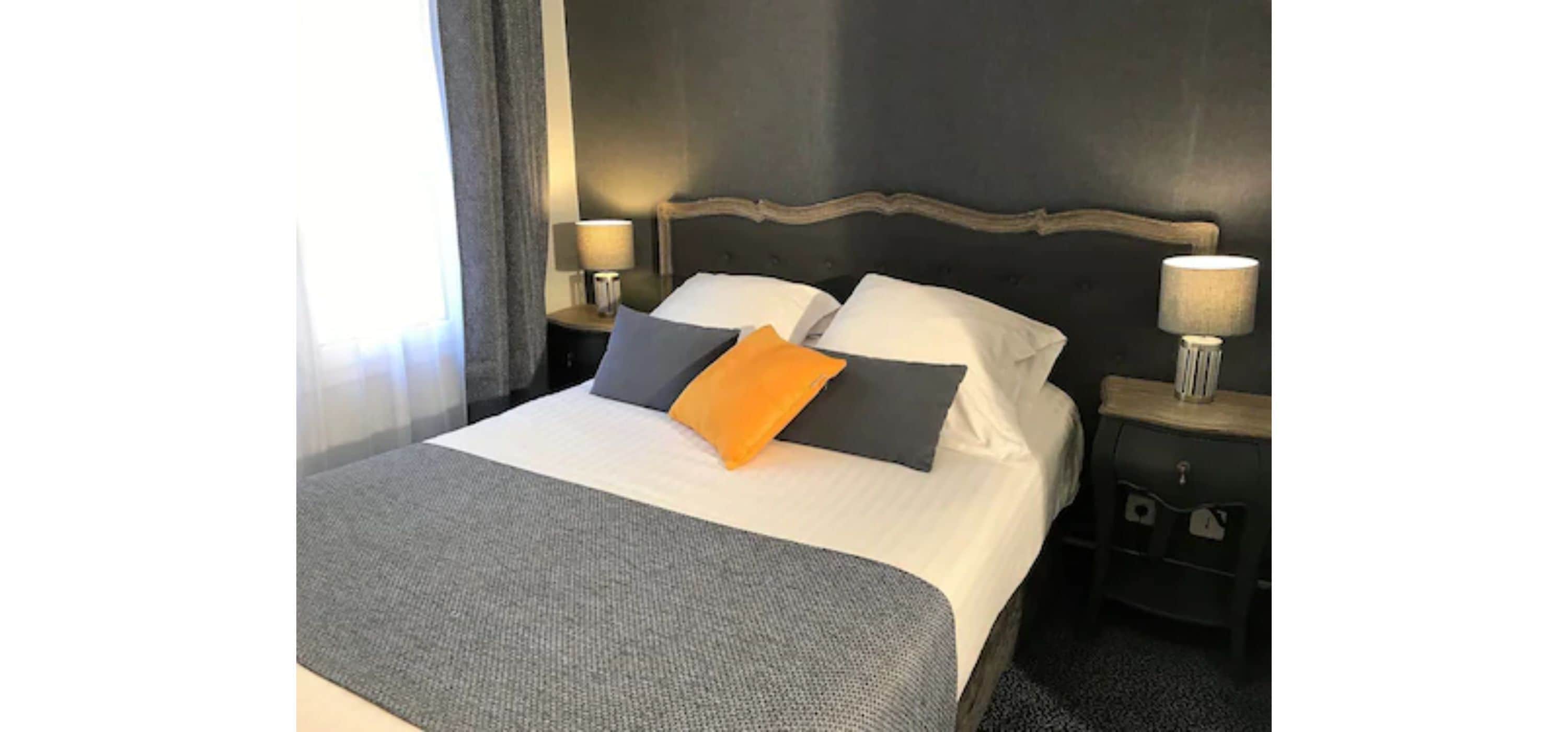 A neatly made bed with a grey blanket, two pillows, and a single orange pillow. There are two bedside lamps on either side of the bed.