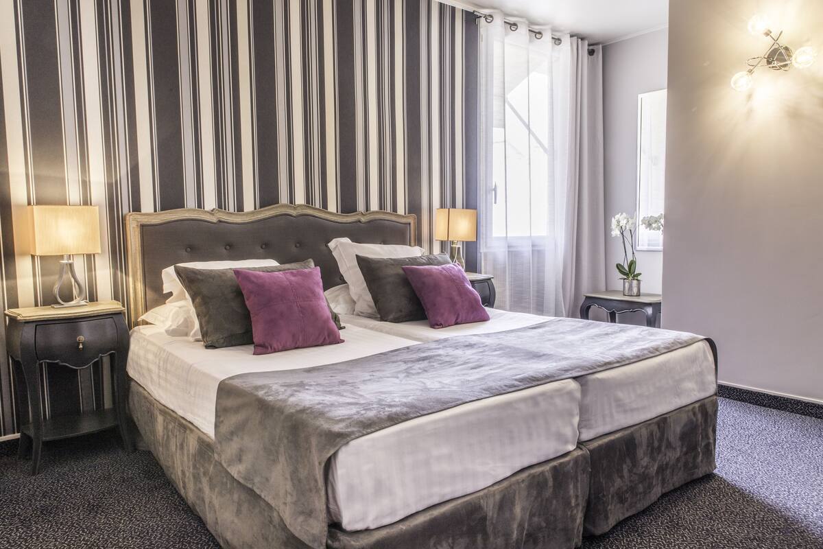 A double bed with a grey headboard, two bedside tables with lamps, a potted plant, and a window with curtains.