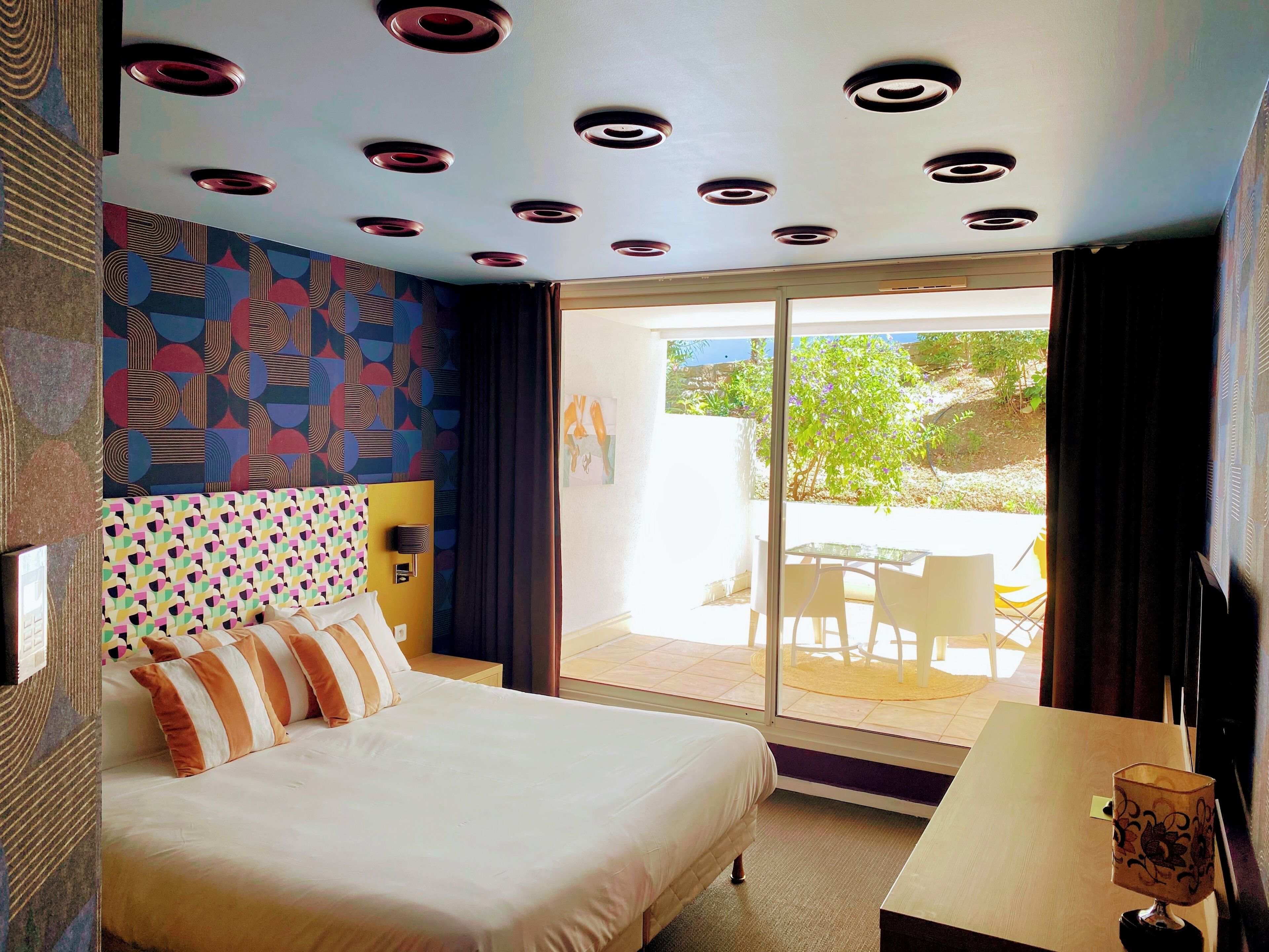 Habitación de hotel con cama, escritorio, cabecero estampado, techo con luces circulares y vista al exterior.