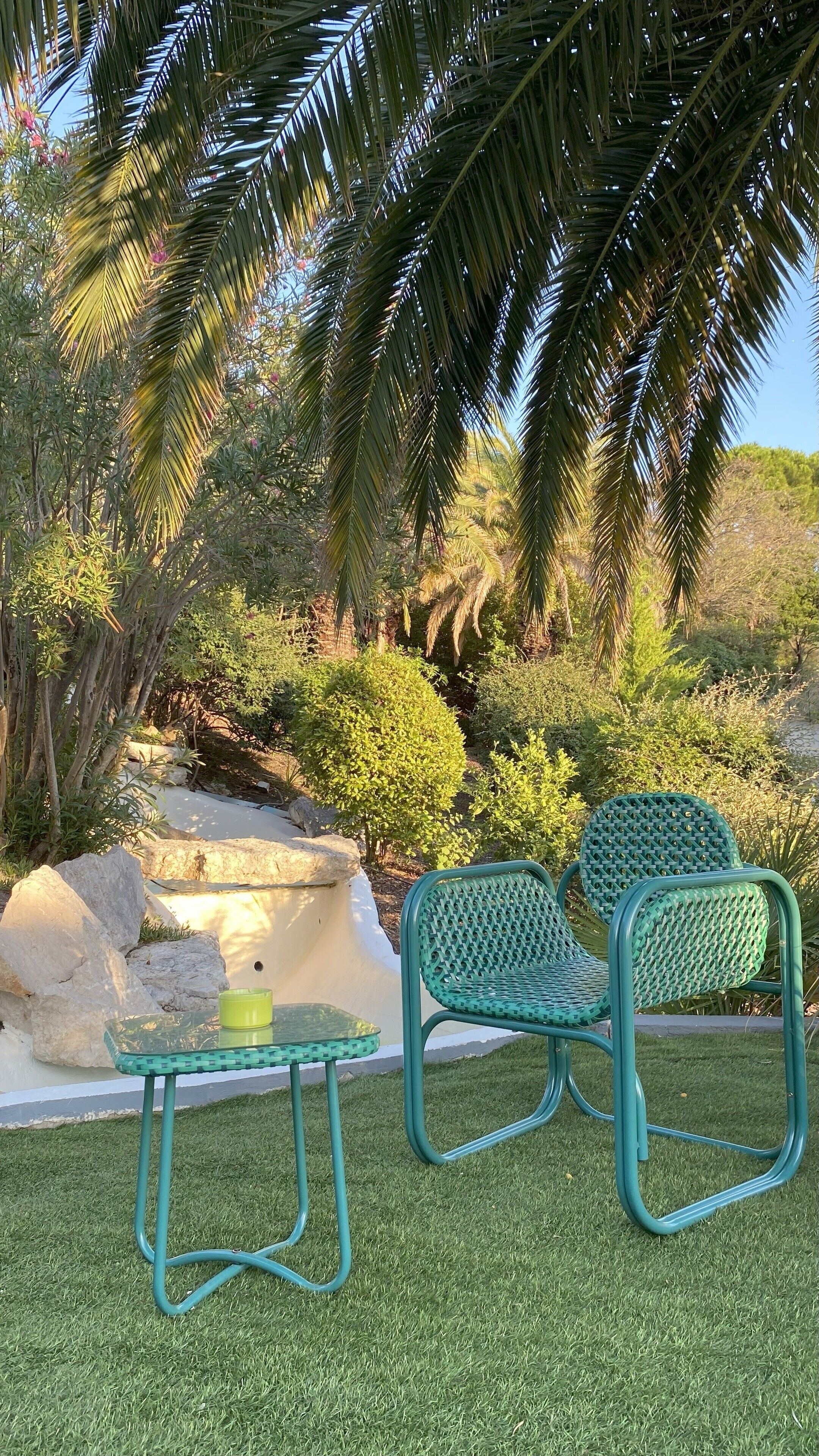 A patio area with a small table and two chairs, surrounded by lush greenery and palm trees.