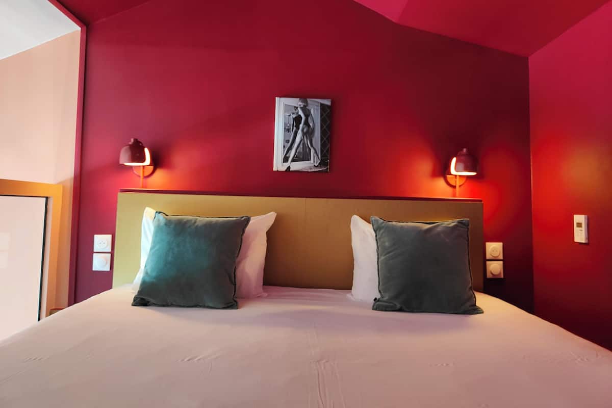 A hotel room with a red wall, a large bed with two pillows, two wall-mounted lamps, and a framed picture on the wall.