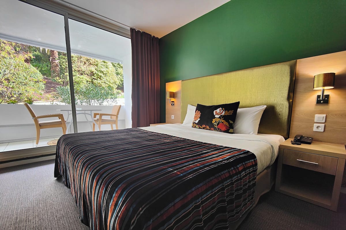 A hotel room with a large bed, a nightstand, a lamp, a chair, and a view of greenery outside.