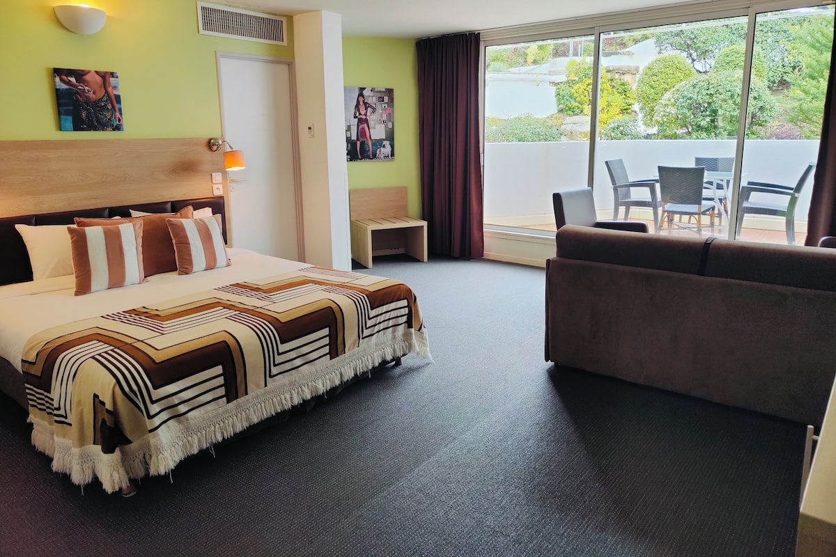 A hotel room with a bed, a sofa, a small table, and a view of the outdoors.