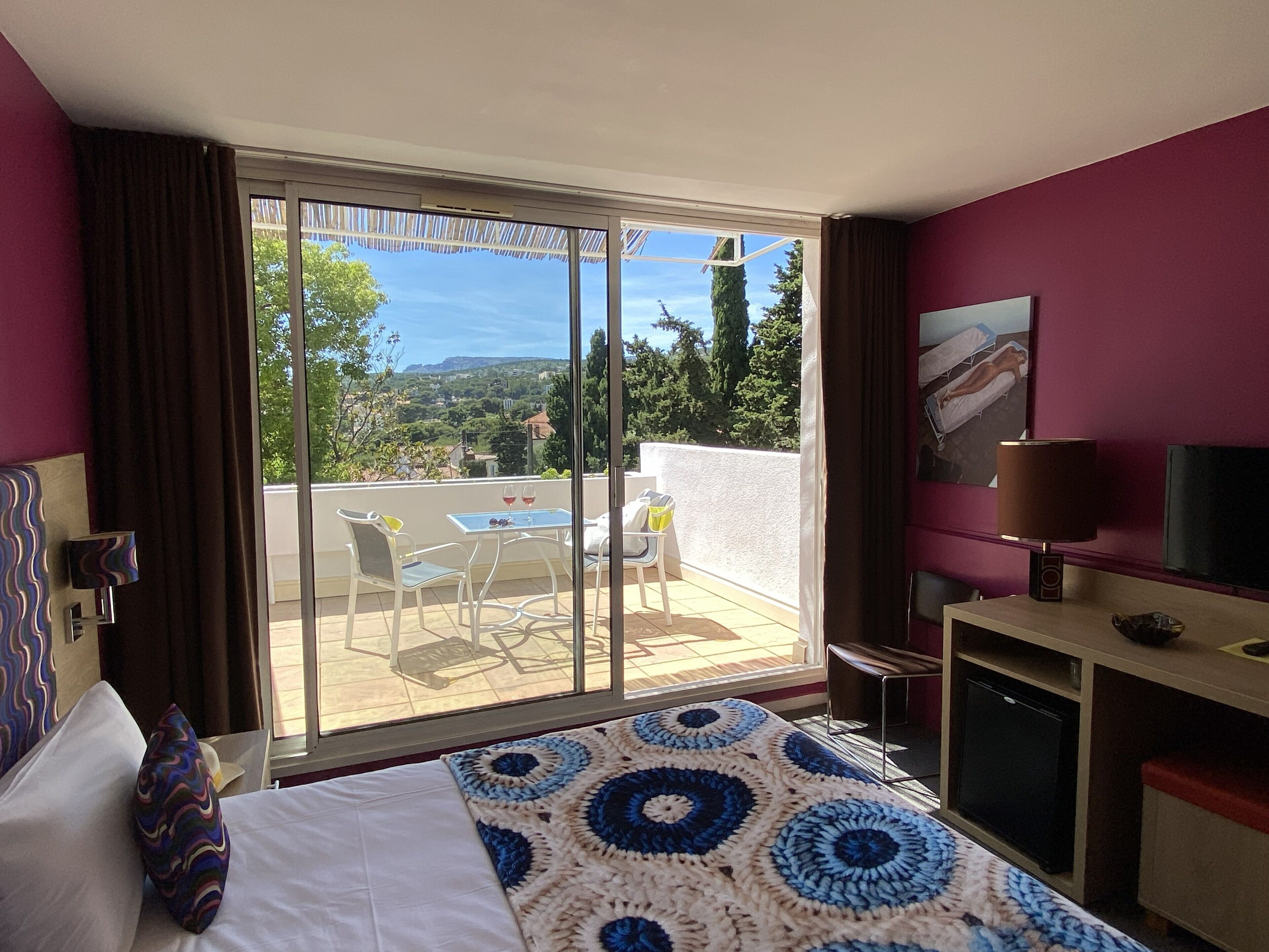 A hotel room with a bed, a desk, a chair, a table with two glasses of wine, and a view of the outdoors.