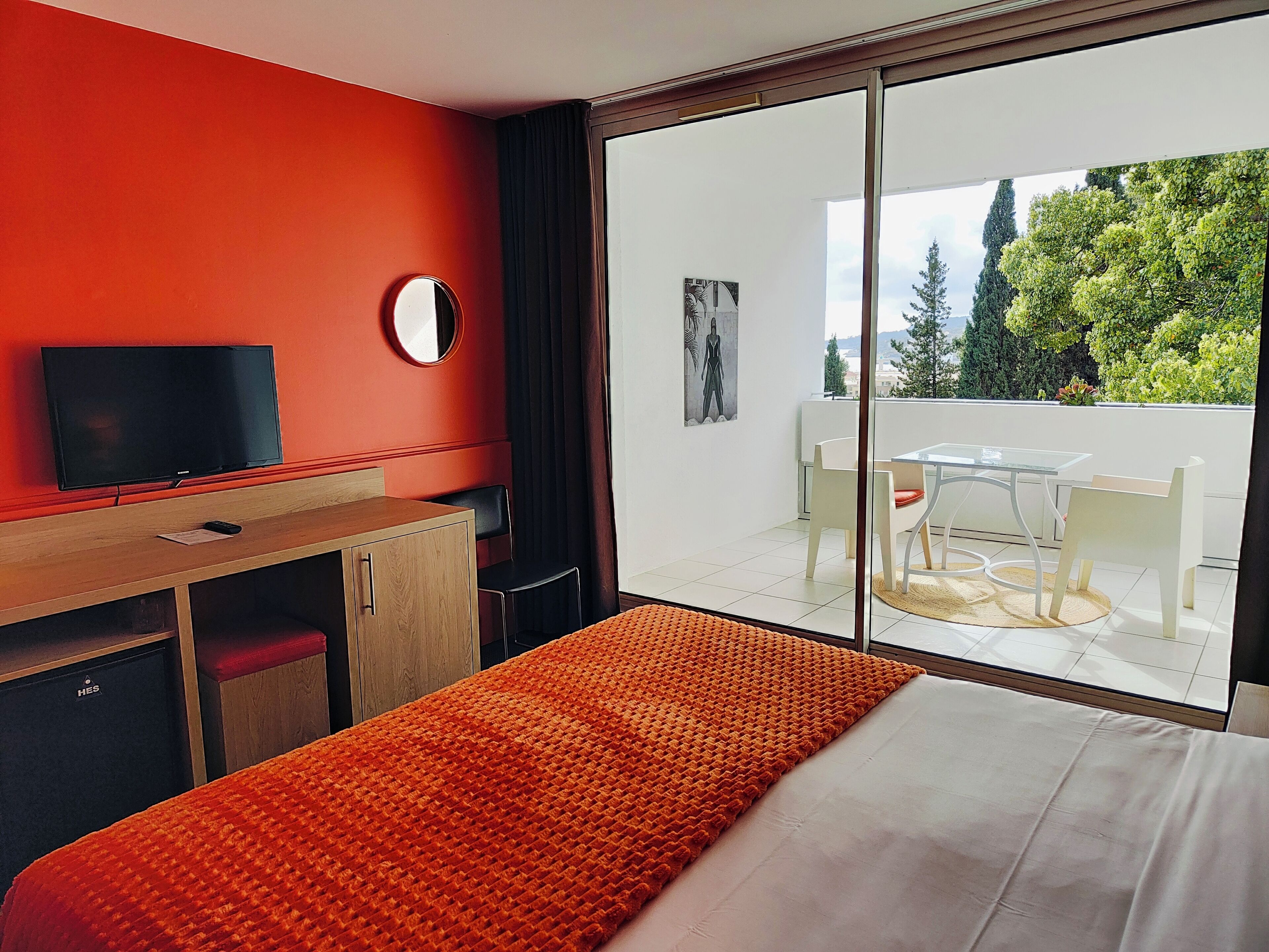 Une chambre d’hôtel comprenant un lit, un bureau avec une télévision, une grande fenêtre donnant sur la verdure et un balcon avec une table et des chaises.