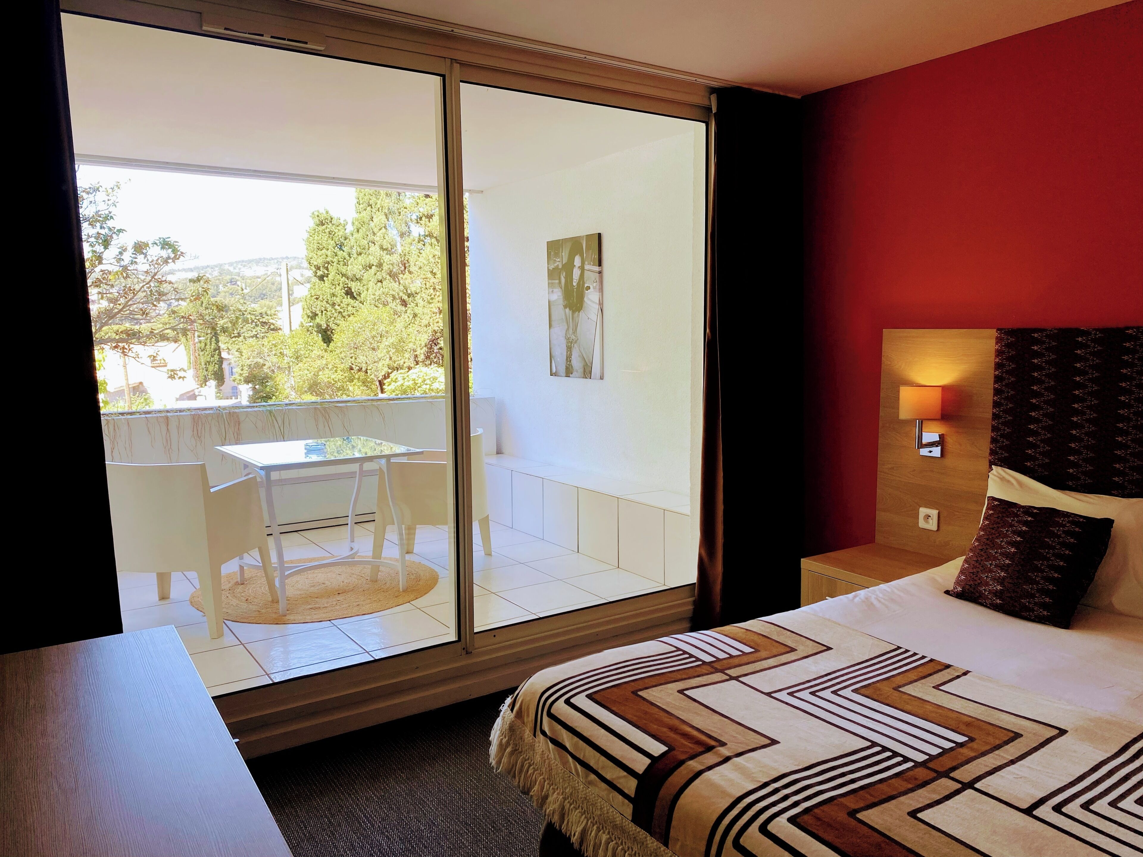 Habitación de hotel con cama, mesita de noche, lámpara, ventana con vistas al exterior y un pequeño balcón con mesa y sillas.