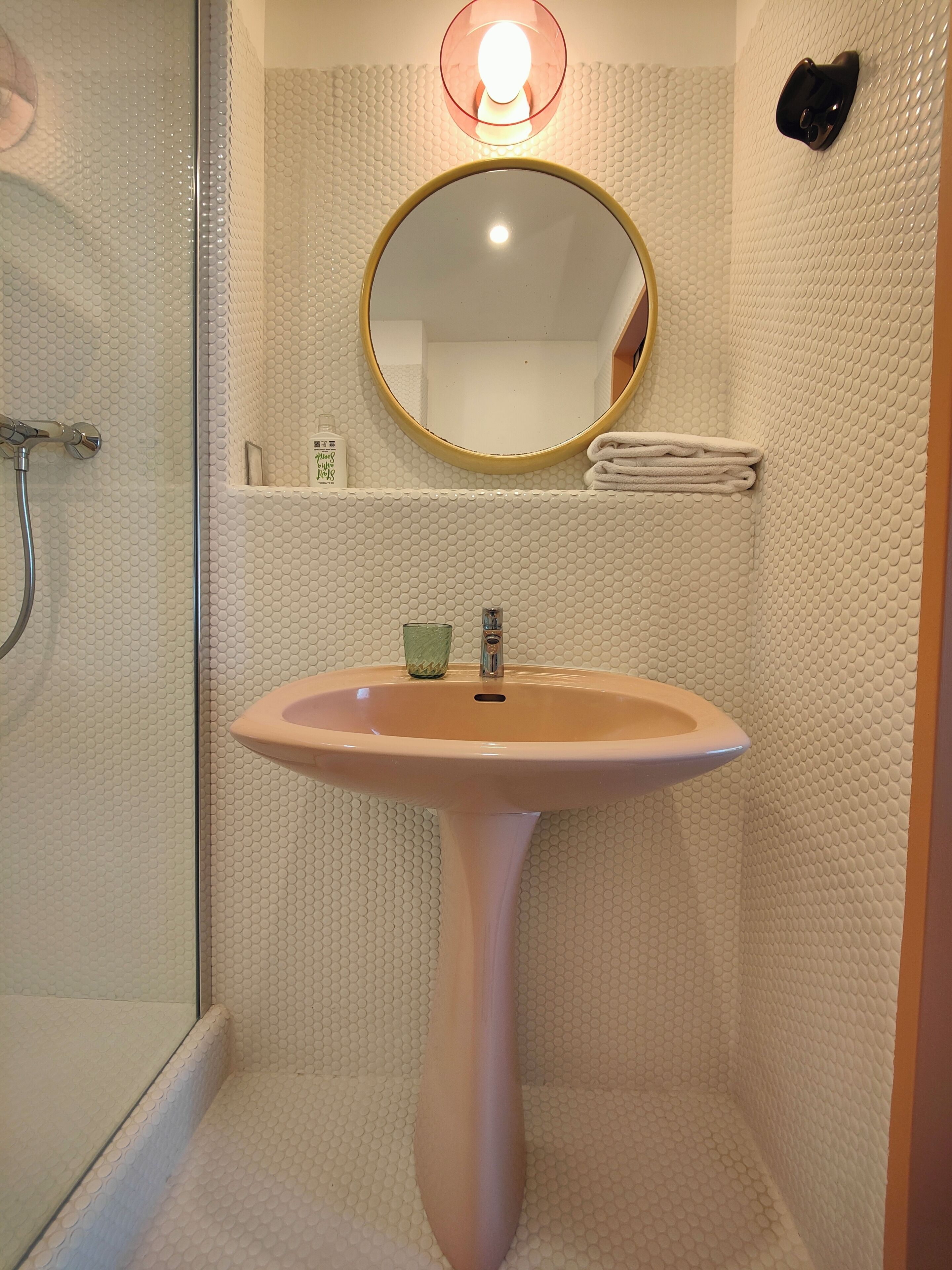 Un bany amb plat de dutxa de vidre, un lavabo pedestal, un mirall rodó i un suport per a tovalloles.