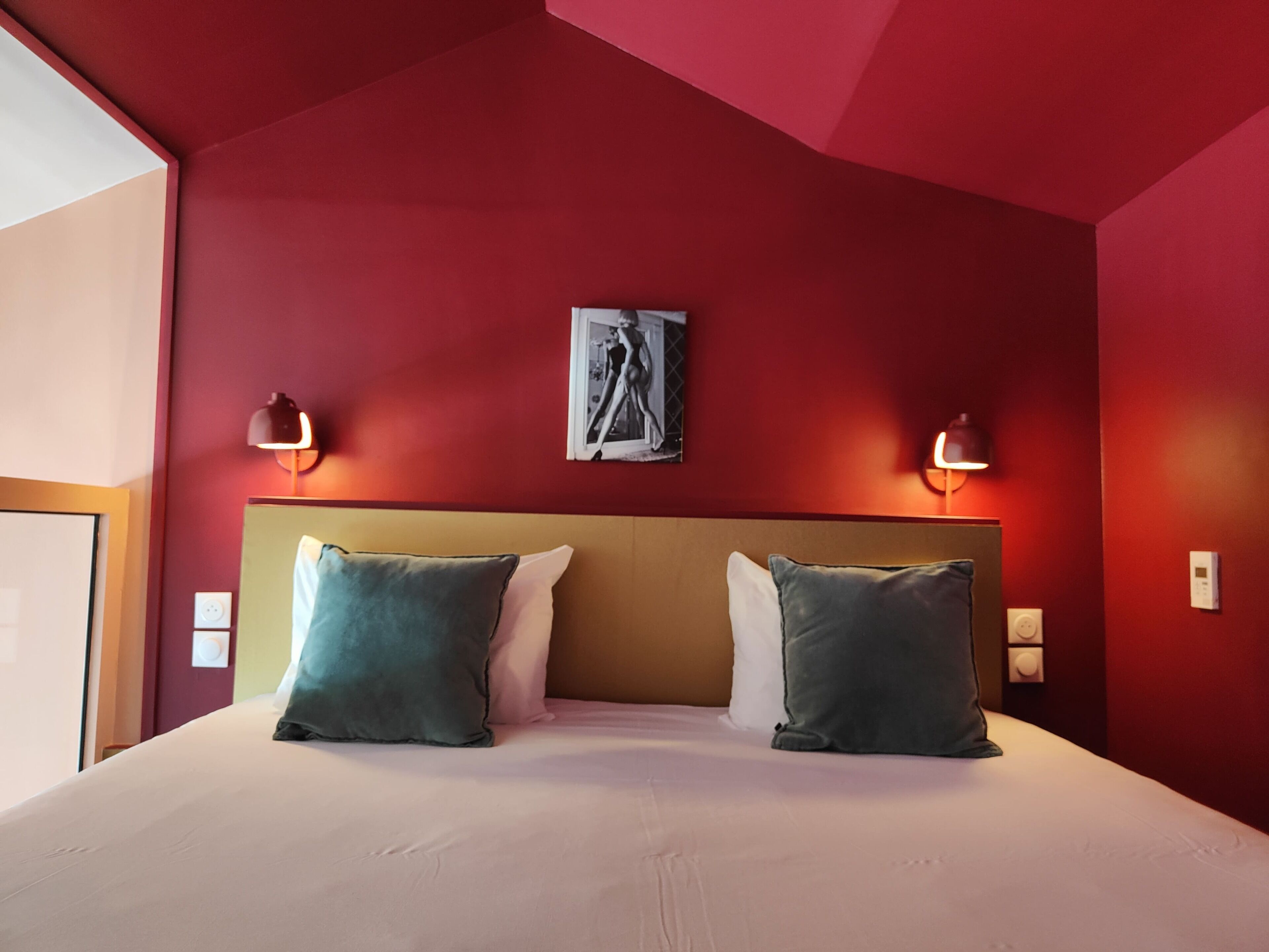 A hotel room with a red wall, a large bed with two pillows, and two wall-mounted lamps.