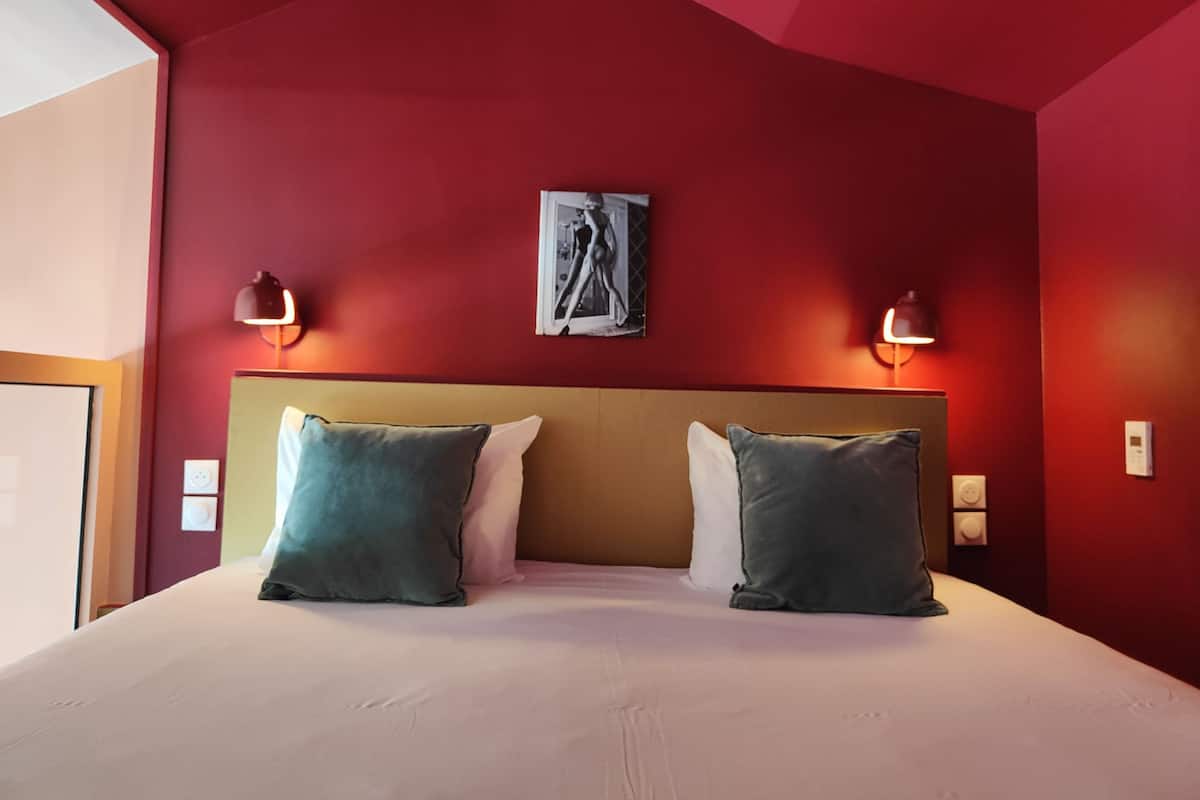 A hotel room with a red wall, a large bed with two pillows, and two wall-mounted lamps.