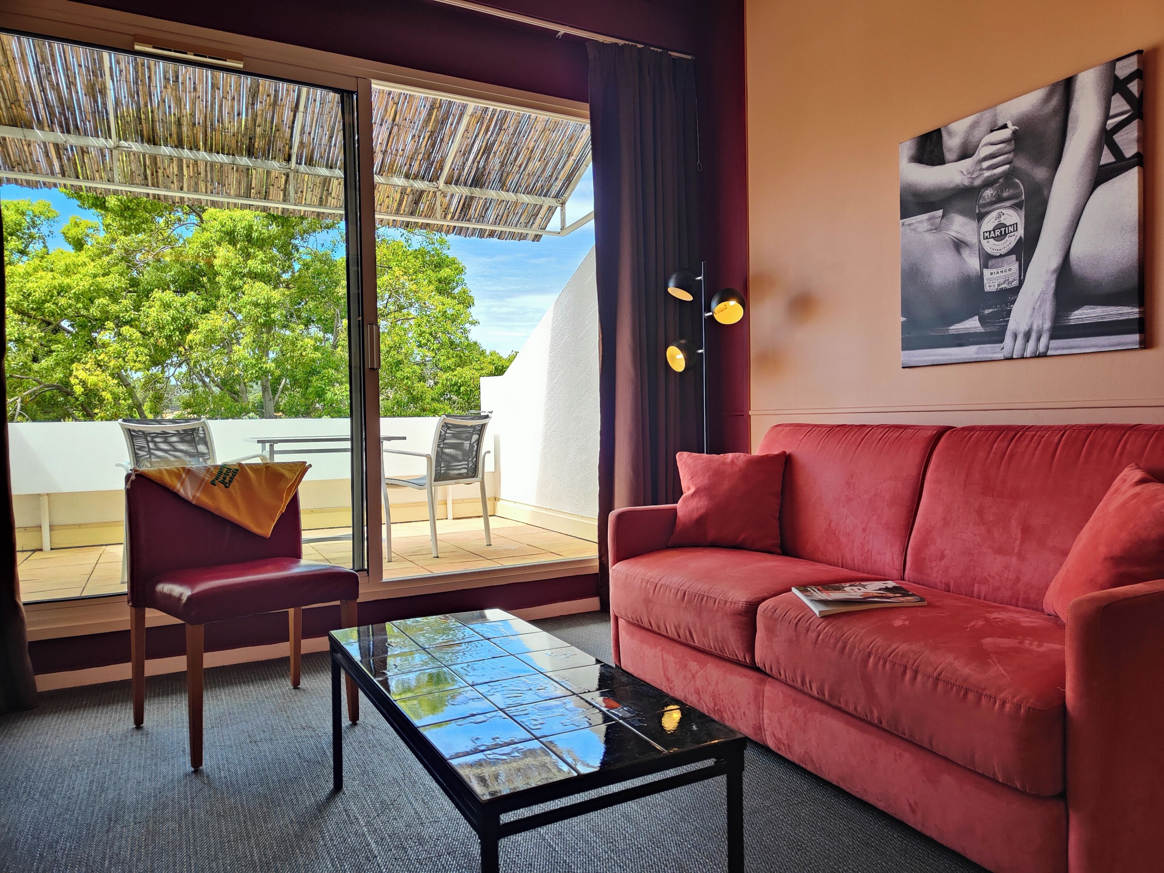 Ein rotes Sofa, ein Glastisch, ein Sessel, ein großes Fenster mit Blick auf Bäume und ein Bild an der Wand.