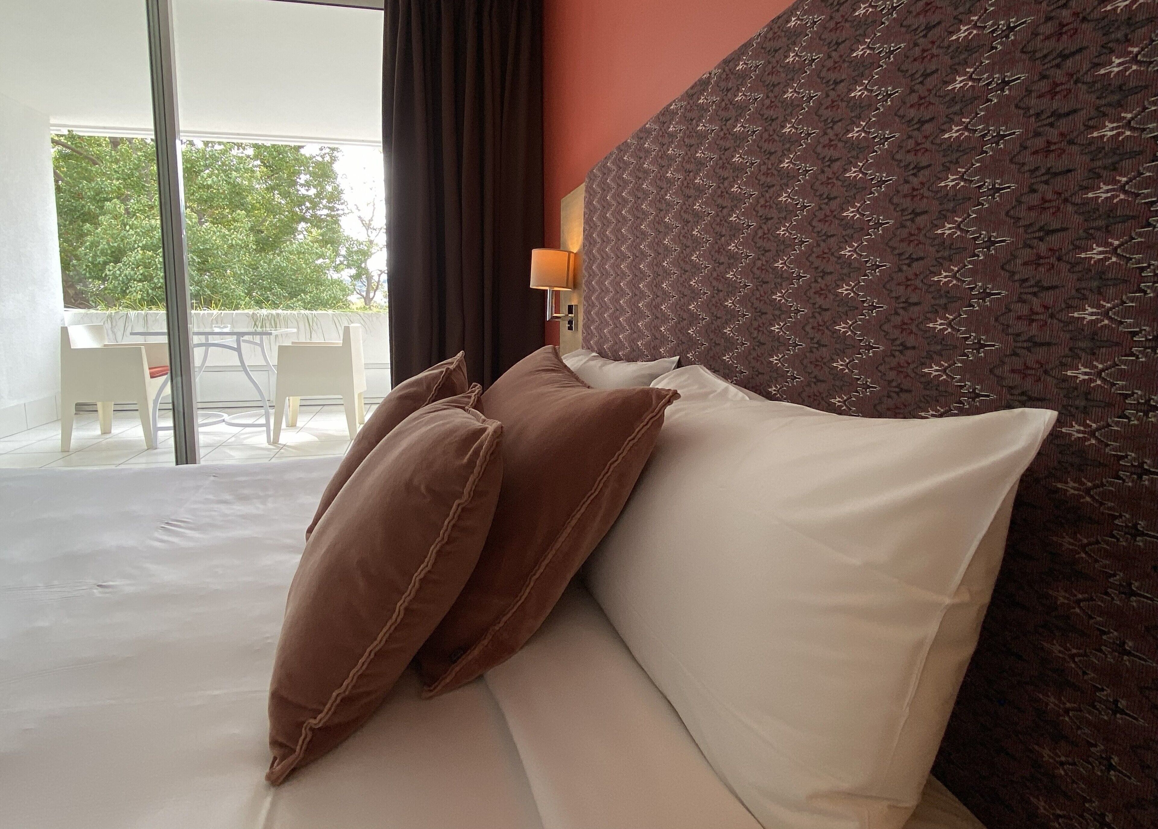 A hotel room with a bed, a headboard, a window with curtains, and a view of the outdoors.