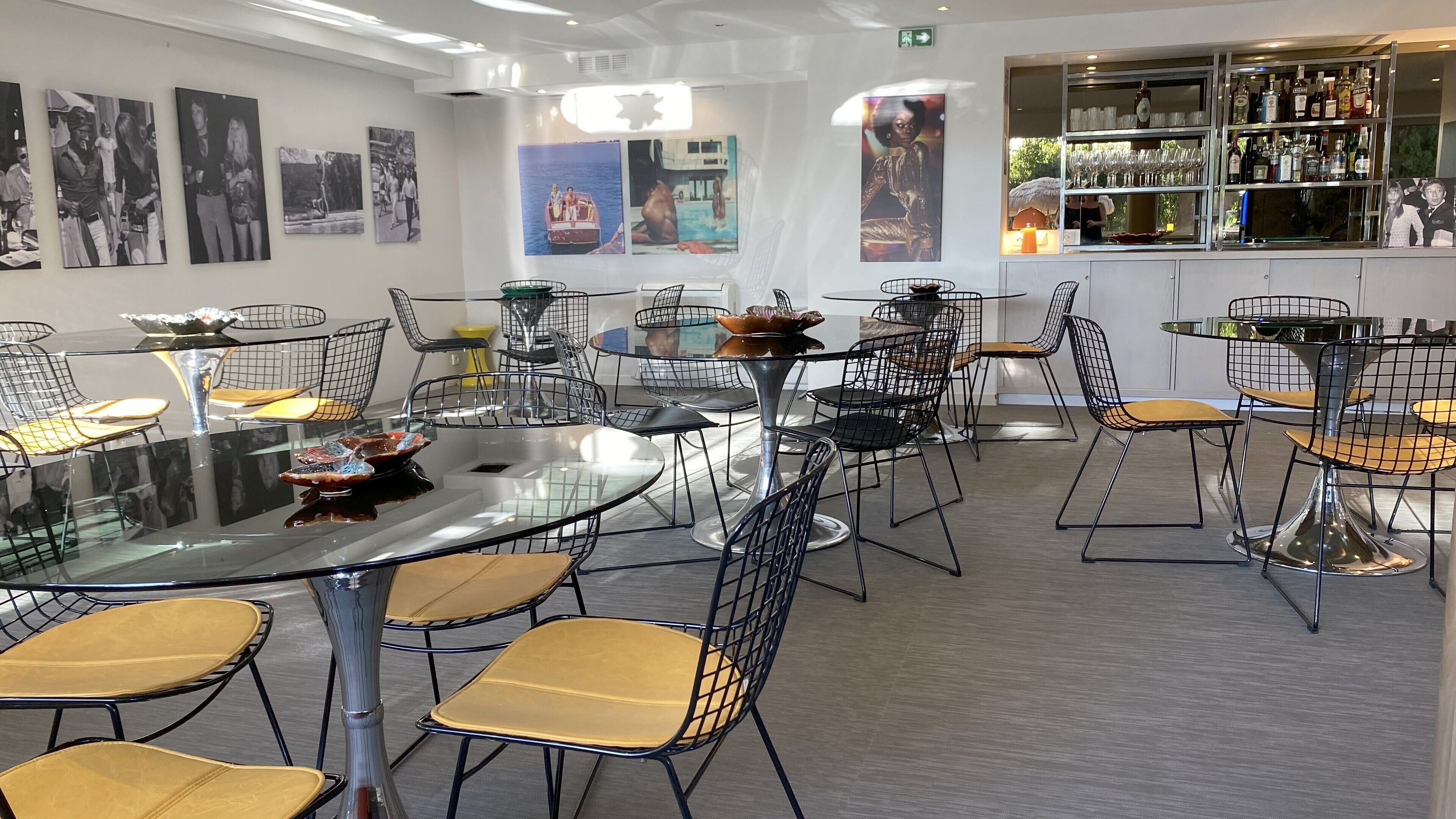 Un comedor moderno con mesas redondas y sillas de malla metálica, con obras de arte en las paredes y una barra con diferentes botellas.