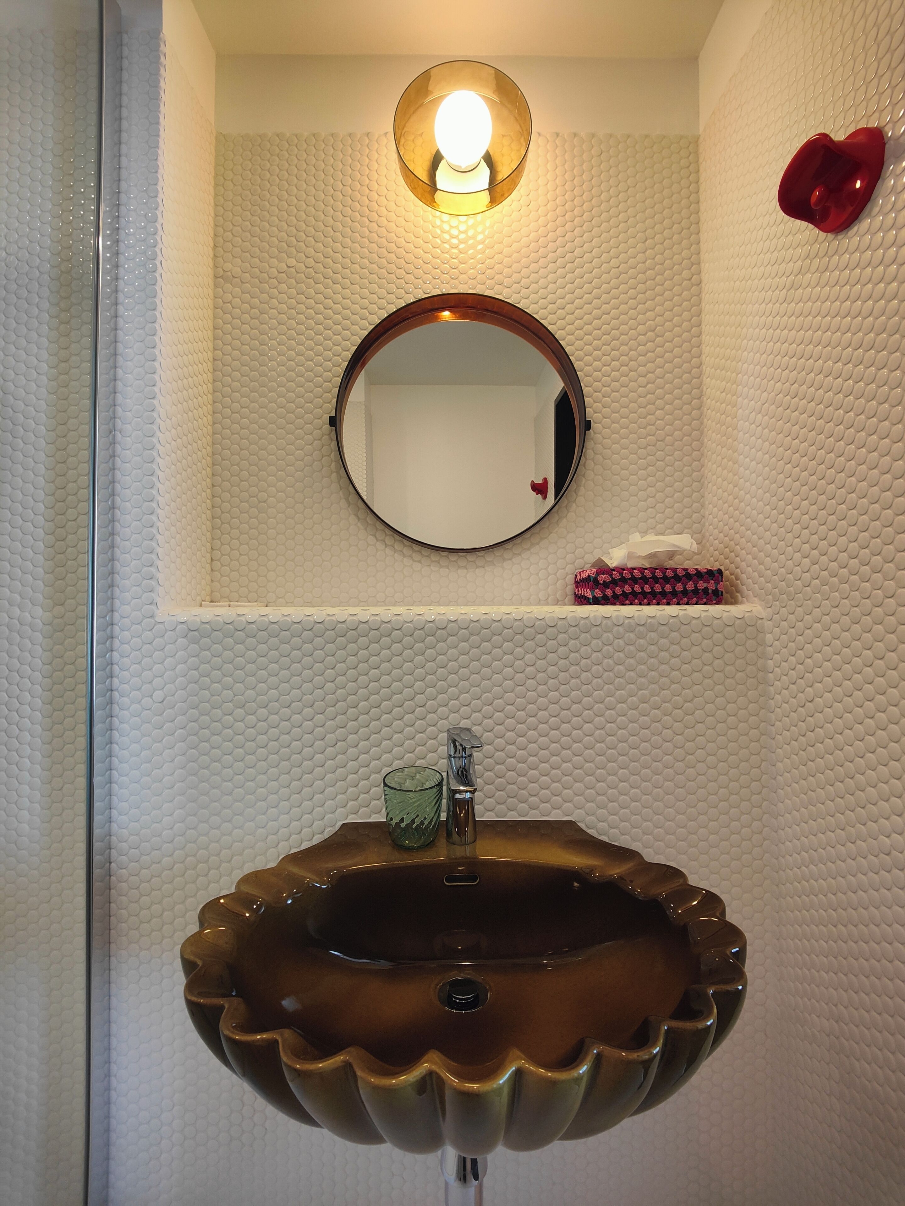 A bathroom with a round mirror, a round sink, a light fixture above the mirror, and a red heart-shaped object on the wall.