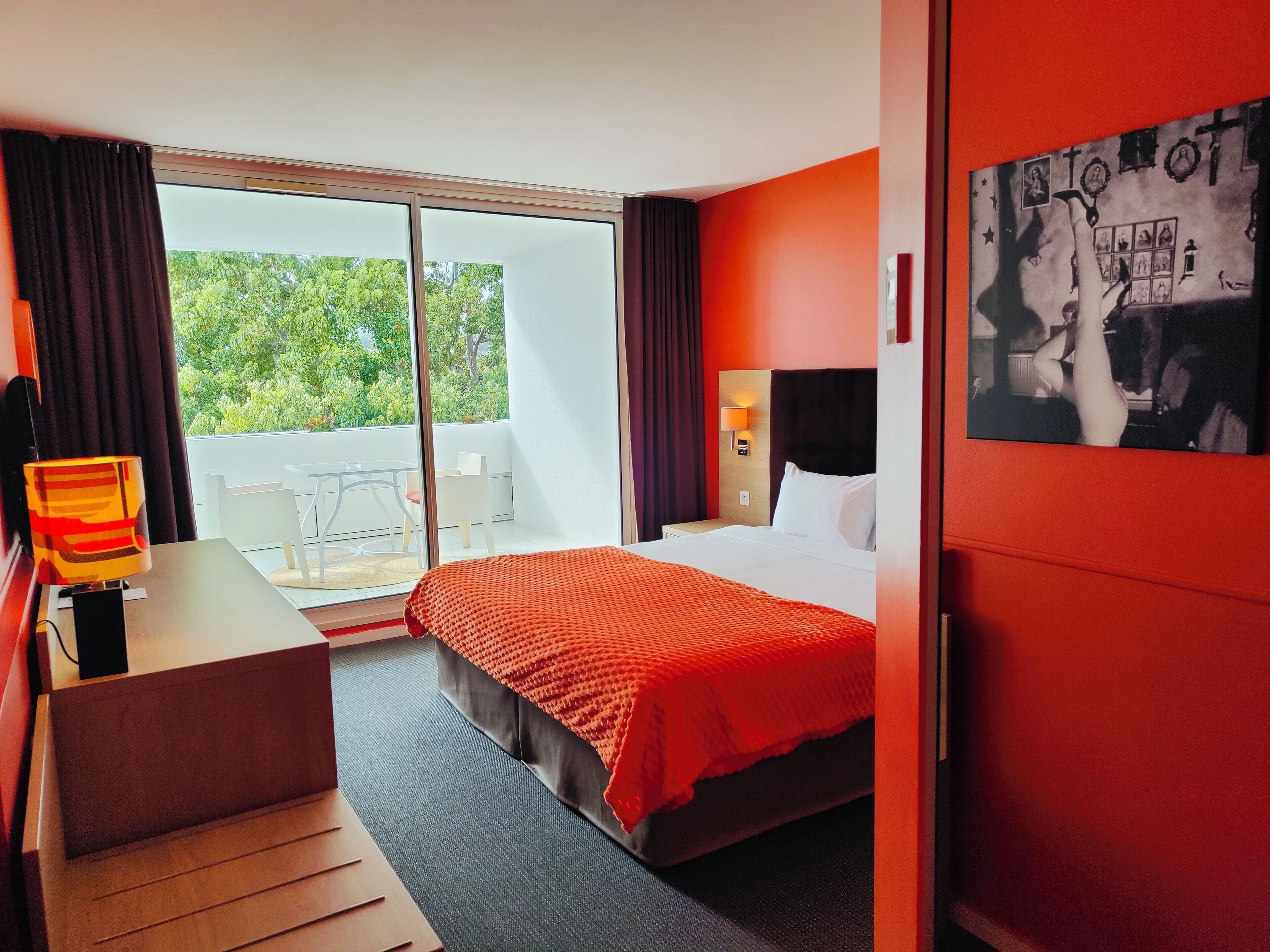 A hotel room with a bed, a desk, a chair, a nightstand, a lamp, a window with curtains, and a wall with a framed picture.