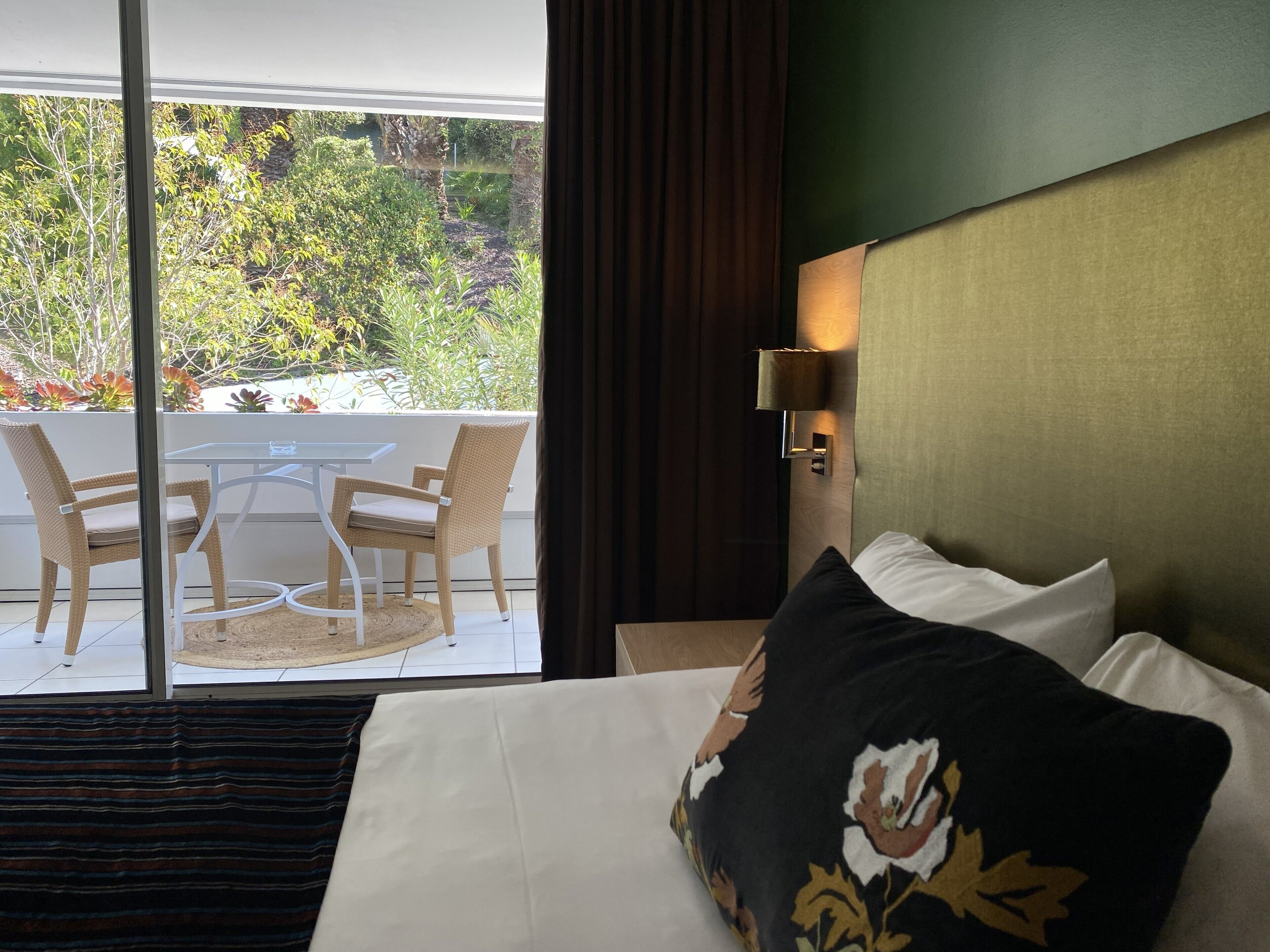 A hotel room with a bed, a table with chairs, a view of a garden, and a wall-mounted lamp.