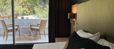 A hotel room with a bed, a table with chairs, a view of a garden, and a wall-mounted lamp.