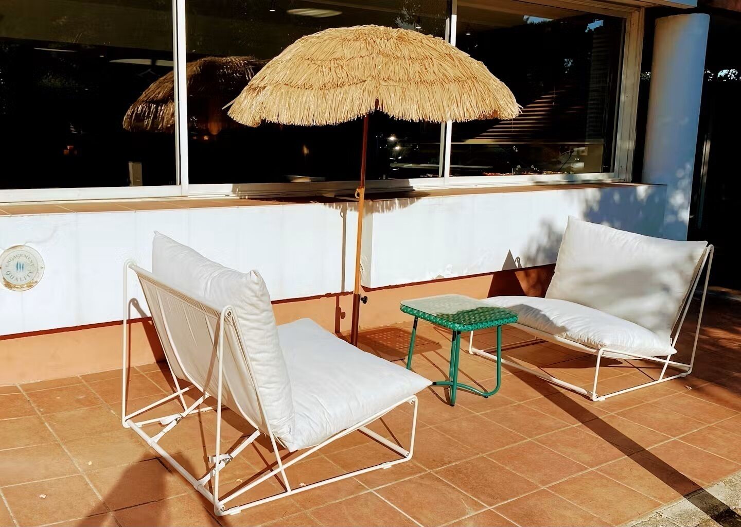 A terrace with two white lounge chairs and a small table, a thatched umbrella, and a view of the city at night.