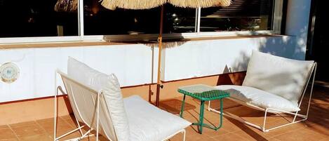A terrace with two white lounge chairs and a small table, a thatched umbrella, and a view of the city at night.