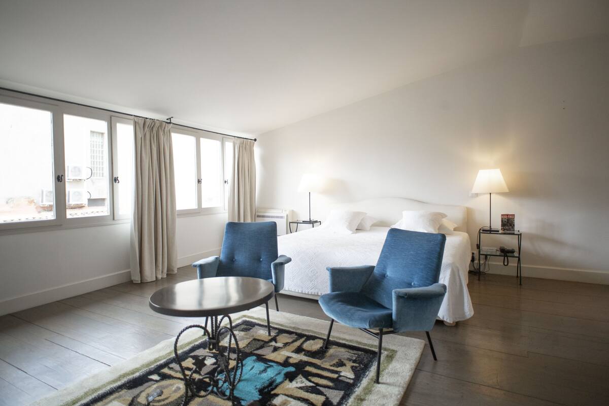 A hotel room with a bed, two blue armchairs, a small round table, a lamp, and a view of the city through large windows.