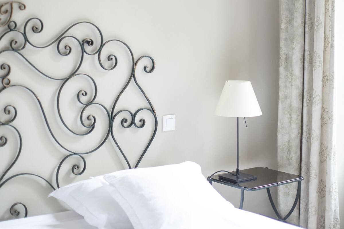 A neatly made bed with white linens, a metal headboard, a bedside table with a lamp, and a wall-mounted light fixture.